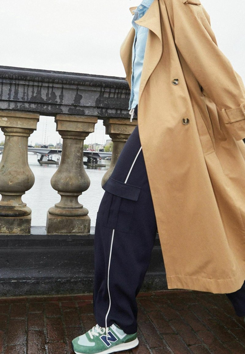 Trench-coat beige, chemise boutonnée bleu clair, pantalons cargo bleu marine avec passepoil blanc, et baskets New Balance vertes, marchant près d'une rivière.