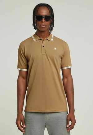 Camisa polo marrón con ribete blanco en el cuello y las mangas, hecha de material suave, combinada con pantalones grises, llevada por una persona con gafas de sol.