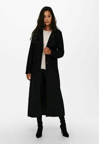 Black long coat made of textured fabric, featuring a notched lapel, front buttons, and side pockets, worn over a light top and dark pants.