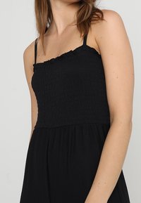 Žena v bezramínkových černých šatech s tenkými ramínky a texturovaným, nabíraným živůtkem na hladkém světlém pozadí.