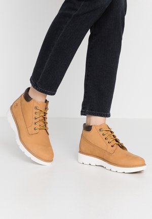 Botas de tobillo de cuero marrón claro con cuello marrón oscuro y cordones amarillos, usadas con unos jeans negros cortos sobre un suelo y un fondo blancos.