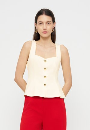 Kvinde med glat, mørkt hår iført en ærmeløs cremefarvet top med knapper og røde bukser, stående med hænderne bag ryggen.