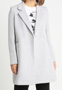 Cappotto grigio chiaro, doppiopetto, con colletto a risvolto, tasche laterali e tessuto strutturato, indossato sopra una maglietta grafica.