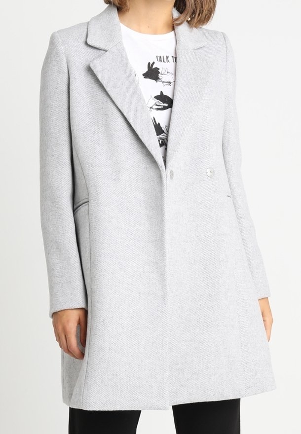 Cappotto grigio chiaro, doppiopetto, con colletto a risvolto, tasche laterali e tessuto strutturato, indossato sopra una maglietta grafica.
