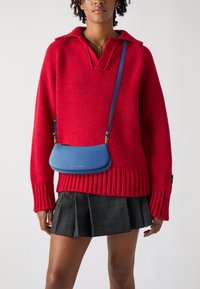 Pulover oversized roșu din tricot, cu decolteu în V și manșete ribbate, asociat cu o geantă din piele albastră tip crossbody și o fustă plisată ecosez cu carouri negre.