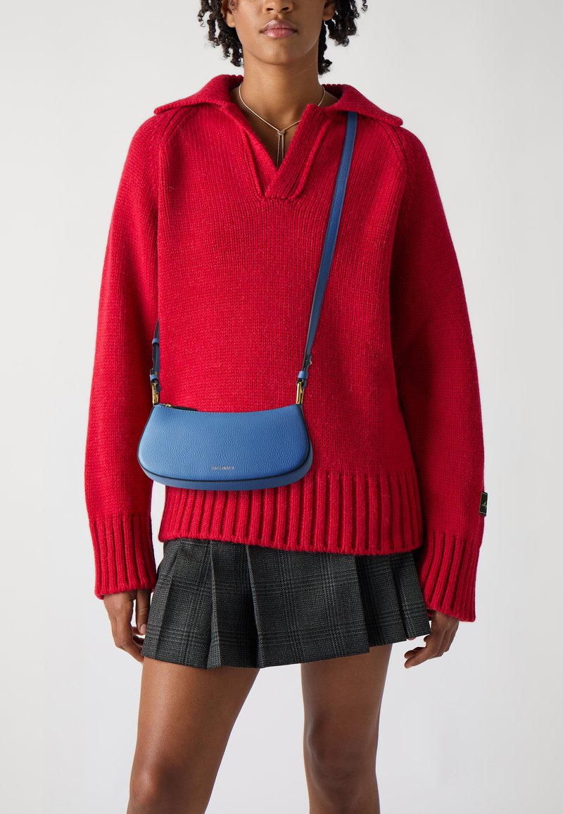 Pulover oversized roșu din tricot, cu decolteu în V și manșete ribbate, asociat cu o geantă din piele albastră tip crossbody și o fustă plisată ecosez cu carouri negre.