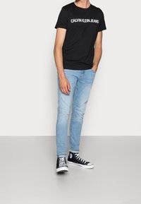 Muž v černém tričku Calvin Klein Jeans, světle modrých úzkých džínách a černých kotníkových teniskách stojí s jednou rukou v kapse.