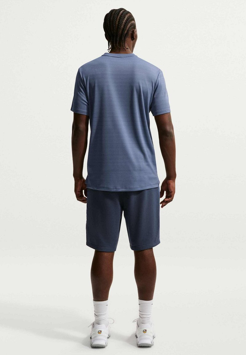 Hombre con el pelo trenzado de espaldas, lleva una camisa azul de manga corta, pantalones cortos azul marino, calcetines blancos y zapatillas deportivas blancas.