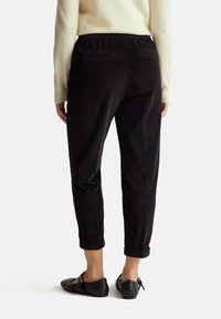 Zwarte corduroy broek met een relaxte pasvorm, elastische tailleband en opgerolde pijpen. Achterzakken zijn zichtbaar. Gedragen met zwarte platte schoenen.