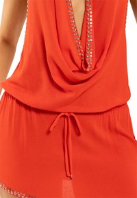 Red sleeveless dress with a deep V neckline adorned with silver studs and a waist tie, worn by a person shown from shoulders to hips.