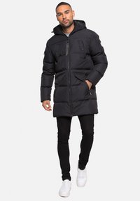 Černá prošívaná bunda s zipem vpředu, velkými kapsami a kapucí. Má prošívanou strukturu a elastické manžety. Je nošena s černými kalhotami a bílými teniskami.