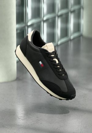 Schwarzer und beiger Sneaker mit Schnürsenkeln, der auf der Seite ein kleines rotes, weißes und blaues Logo zeigt, vor einem grauen Boden und einer Glaswand.