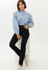 Hellbluse mit Streifen, hohen Kragen und Puffärmeln, kombiniert mit schwarzen Skinny Jeans und weißen Sneakers, vor einem neutralen Hintergrund.