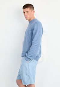 Maglione azzurro chiaro a maglia con texture a coste, collo rotondo e spalle scese, abbinato a pantaloni corti azzurri chiari. Borsa tote bianca semplice.