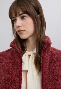 Chaqueta de forro polar roja con cuello alto y cierre de cremallera, que presenta una superficie texturizada. Llevada sobre una blusa de color claro con un detalle de lazo.