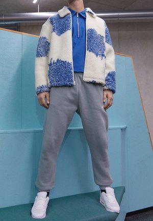 Fleecejacke mit blau-weißen Mustern, kombiniert mit einem blauen Polo-Shirt und grauen Sweatpants, getragen mit weißen Sneakers.
