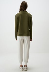 Felpa verde oliva con colletto a risvolto, abbinata a pantaloni bianchi a sigaretta e scarpe da ginnastica chiare, vista dal retro.