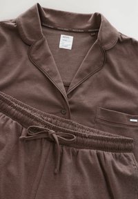 Ruskea pyjamapuku, joka on valmistettu pehmeästä kankaasta, sisältää kauluksellisen paidan, jossa on nappi, ja joustovyötäröiset housut, joissa on naru.