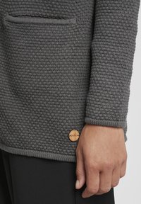 Gray knitted cardigan with a textured pattern, featuring a small wooden logo tag and a front pocket. The fabric appears thick and warm.