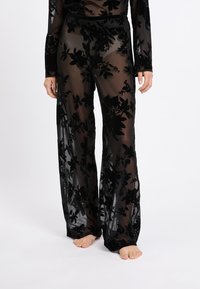 Schwarze durchsichtige Hosen mit floralem Samtüberzug, weit geschnitten, elastischem Bund und langen Ärmeln, die ein schichtartiges, strukturiertes Aussehen erzeugen.