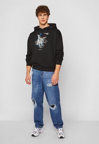 Sudadera negra con un diseño gráfico en azul y plata, combinada con jeans azules desgastados. Modelo que lleva zapatillas blancas y grises.