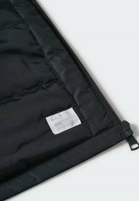 Sort, kviltet stofpose med et hvidt mærke til navn og telefonnummer. Indeholder en lynlås samt teksturerede detaljer langs kanterne.