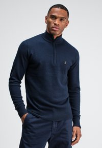 Marineblauwe gebreide pullover met een hoge kraag en een kwart-rits. Heeft een gestructureerde stof en een klein merkembleem op de borst. Strak aansluitende pasvorm.