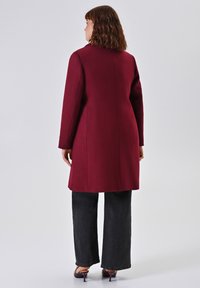 Vrouw met schouderlang bruin haar, gekleed in een lange bordeauxrode jas, zwarte wijde broek en bordeauxrode hakken, staand met de rug naar ons gekeerd.