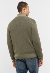 Olivgrön ribbad stickad tröja med hög krage, med texturerade vertikala ränder och åtsittande ärmslut, tillsammans med blå jeans.