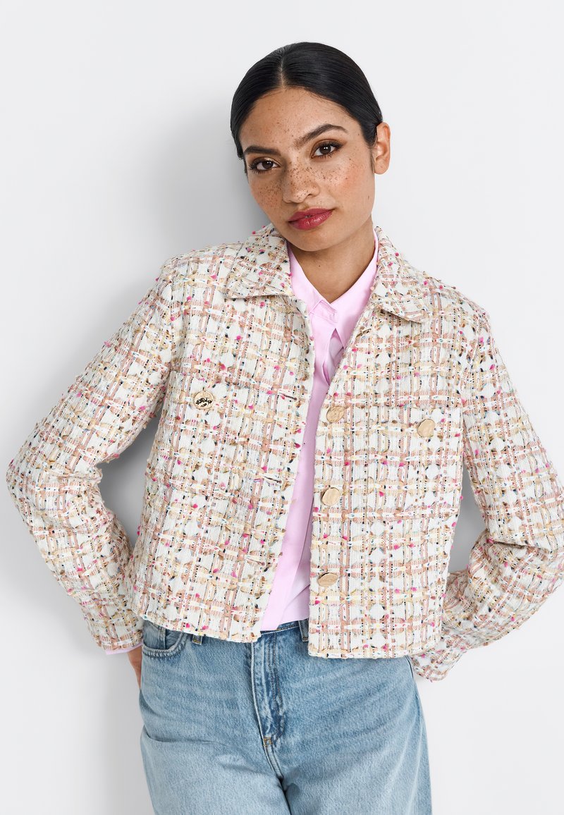 Giacca corta in tweed crema con macchie multicolori, due tasche frontali e grandi bottoni, indossata sopra una maglietta rosa chiaro e jeans blu.