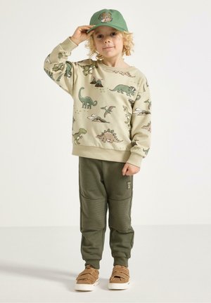 Beżowa bluza z nadrukiem dinozaura, zielone spodnie z pikowanymi akcentami i brązowe buty na rzepy; zielona czapka z motywem zwierzęcym.