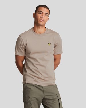 Bărbat îmbrăcat cu un tricou bej cu mânecă scurtă și un logo galben, purtând pantaloni cargo verde măsliniu, stând cu mâinile la spate.