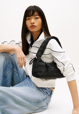 Młoda kobieta z długimi ciemnymi włosami siedzi, ubrana w białą kurtkę z czarnymi paskami, jasnoniebieskie dżinsy oraz czarną torebkę na ramię w wzory.