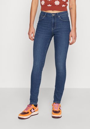 Personne portant un jean skinny bleu foncé, un crop top orange à fleurs et des baskets orange et violettes, debout sur un sol gris clair.