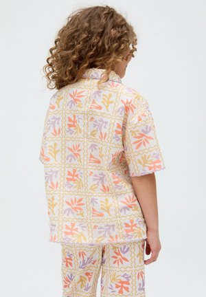 Die Person mit lockigem Haar, die von hinten zu sehen ist, trägt ein passendes kurzärmeliges Hemd und Shorts mit gelbem, orangefarbenem und lila Blatt-geometrischem Muster.