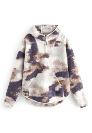 Personne vêtue d'un pull polaire tie-dye à zip quart, de leggings côtelés beige, de chaussettes blanches et de baskets blanches New Balance sur un fond uni.