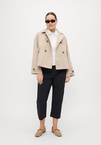 Beige kort jakke med dobbeltknap, hvit skjorte, svarte capribukser og lysebrune slip-on sko. Solbriller fullfører antrekket.