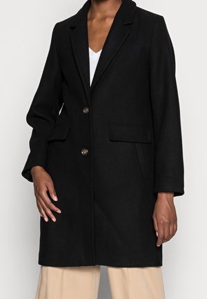 Personne portant un manteau noir boutonné avec des poches à rabat sur un top blanc et un pantalon beige, debout devant un fond clair uni.
