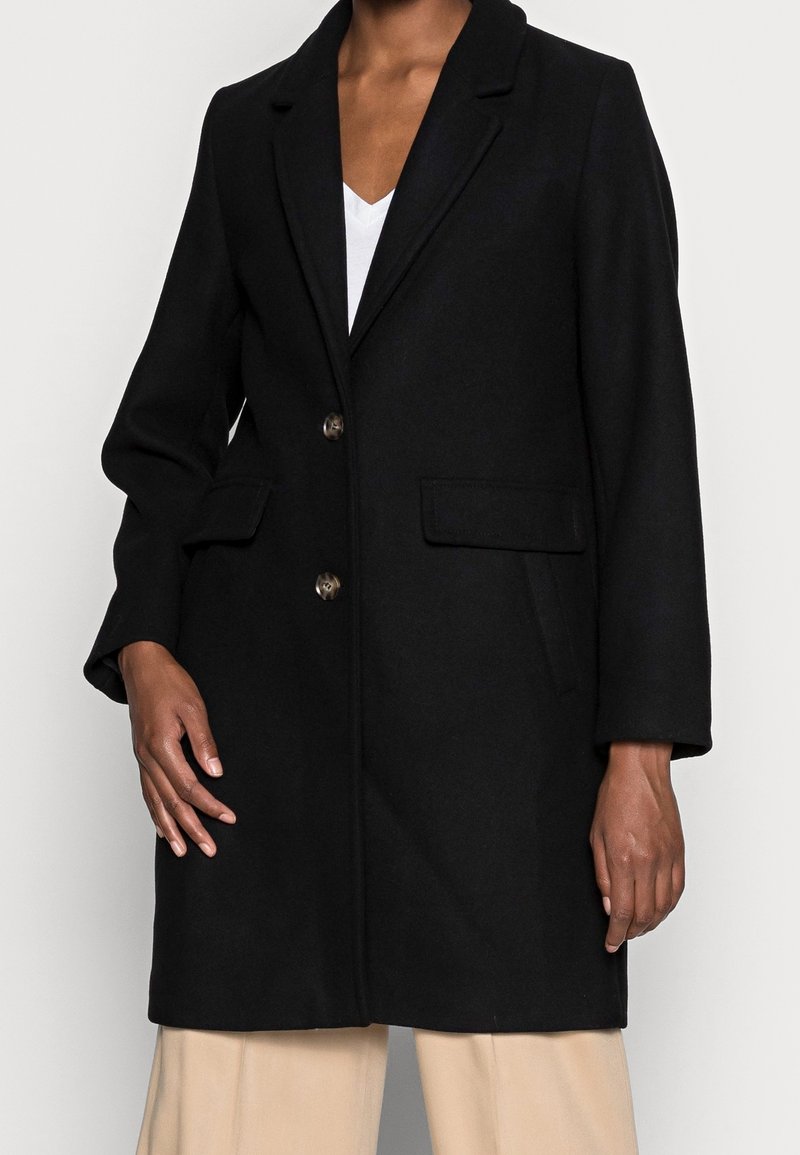 Personne portant un manteau noir boutonné avec des poches à rabat sur un top blanc et un pantalon beige, debout devant un fond clair uni.