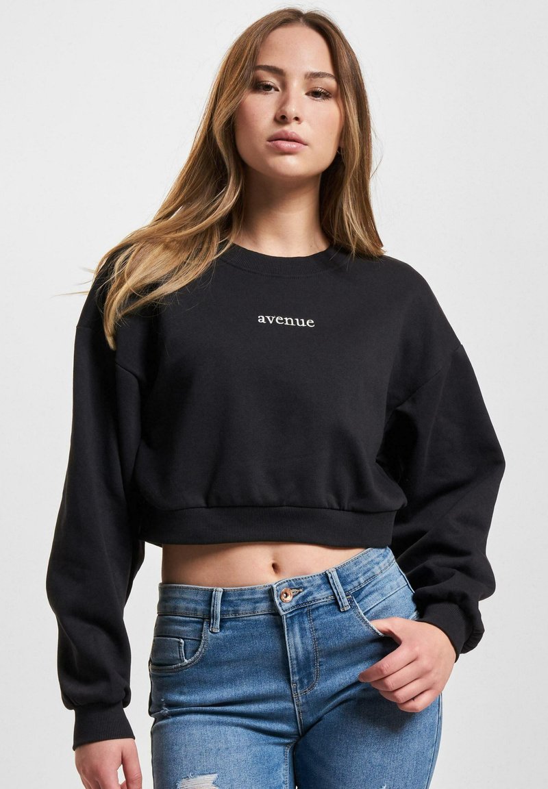 Svart cropped sweatshirt med rund hals og avslappede ermer, med hvit "avenue" tekst på fronten, kombinert med blå denim jeans.