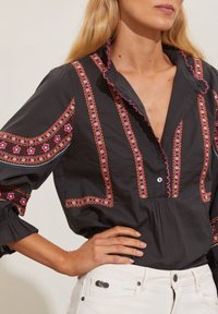 Chemise noire brodée avec des motifs floraux colorés, col à volants et devant boutonné ; poignets conçus avec un détail élastique.