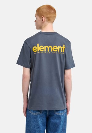 Jeune personne aux cheveux rouges courts porte un T-shirt gris foncé avec un grand texte jaune "element" et un jean bleu, debout de dos.