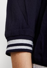 Tessuto blu navy con texture e maniche corte a coste, caratterizzato da strisce orizzontali argento e bianche sul polsino.