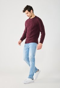 Bordo pleteni pulover s uzorkom pletenice, uparen s svijetloplavim trapericama i bijelim tenisicama. Pulover ima rebraste rukave i donji rub.