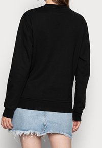 Sudadera negra de manga larga, cuello redondo, con puños y dobladillo de canalé. Lleva puesta una falda de mezclilla destrozada en azul claro.