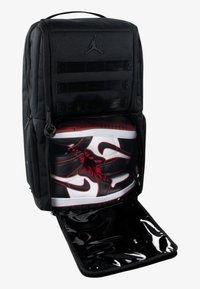 Mochila negra con textura hexagonal, que presenta un gran compartimento frontal que contiene zapatillas Nike rojas y negras. Apertura con cremallera visible.