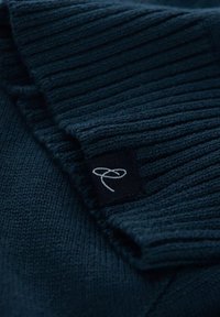 Tejido de suéter de punto azul marino con cuello acanalado. Pequeña etiqueta negra con logotipo blanco cosida en el lateral. Textura suave y patrón detallado.