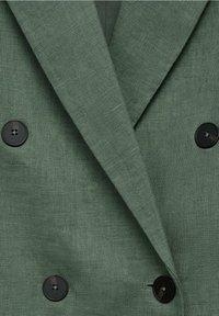 Blazer verde de doble botonadura hecho de un tejido texturizado, con botones negros y solapas con muesca. Primer plano del material y los detalles del diseño.