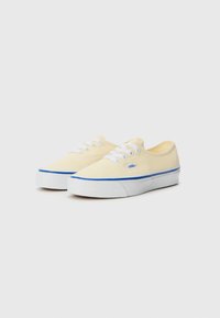 Canvas sneakers in pale yellow with white laces, a blue stripe along the bottom, and a logo tag on the side. Low-profile design.