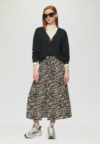 Schwarze gerippte Strickjacke, cremefarbene Rollkragenbluse, gemusterter langer Rock in Schwarz und Beige, silberne Sneakers mit grauen Akzenten, oversized schwarze Sonnenbrille.